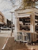 A quaint outdoor espresso bar kiosk with two white stools and a wooden counter sits on a city sidewalk. Above, a sign reads 'espresso BAR kiosk', and in the background, city buildings and street crossings are visible, with cars and a pedestrian present. The scene is partially shaded by overhanging tree branches.