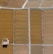 Aerial view of a series of rectangular agricultural fields with varying shades of brown and beige. A solitary wooden structure is situated in one of the fields, casting a shadow on the ground. The fields are divided by narrow pathways or irrigation channels.