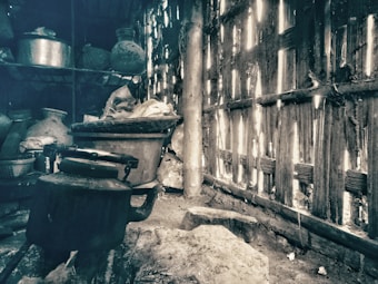 A dimly lit, rustic kitchen with weathered wooden walls allowing slivers of light to enter. Various pots and containers clutter the shelves in the background. A large kettle or pot sits in the foreground on a rough stone floor.