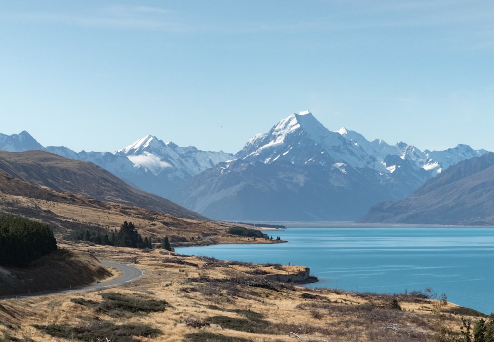 gray road near body of water and snow-capped mountains at daytime