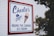 A large sign mounted on a white paneled wall features the text 'Charlie's' prominently in blue. Below it, there is an illustration of an ice cream cone with two scoops, and nearby, the words 'Sherman's Finest Ice Cream' are written in a classic script. The sign includes the phrase 'Round the Corner Ice Cream' in bold letters at the bottom. A red border outlines the sign, and some foliage is visible in the foreground.
