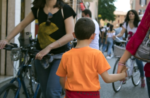 A child wearing an orange shirt is walking and holding hands with an adult in a busy street. People around them are walking and some are riding bicycles. The adults are casually dressed, and the scene has a lively, bustling atmosphere.