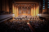 A large orchestra performs on stage in a grand concert hall with wooden walls and a pipe organ backdrop. The hall is filled with an audience seated in rows, and additional spectators are visible in the balcony sections. The conductor stands in the middle, leading the musicians.