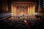 A large orchestra performs on stage in a grand concert hall with wooden walls and a pipe organ backdrop. The hall is filled with an audience seated in rows, and additional spectators are visible in the balcony sections. The conductor stands in the middle, leading the musicians.