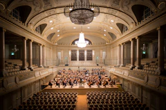 An orchestra on stage in an ornate concert hall with rows of empty seats