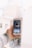 A hand is holding a smartphone with an application open on the screen. The background appears to be a blurred cityscape with tall buildings. The smartphone screen displays profile information and various statistics relating to the profile.
