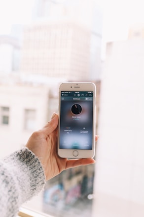 A hand is holding a smartphone with an application open on the screen. The background appears to be a blurred cityscape with tall buildings. The smartphone screen displays profile information and various statistics relating to the profile.