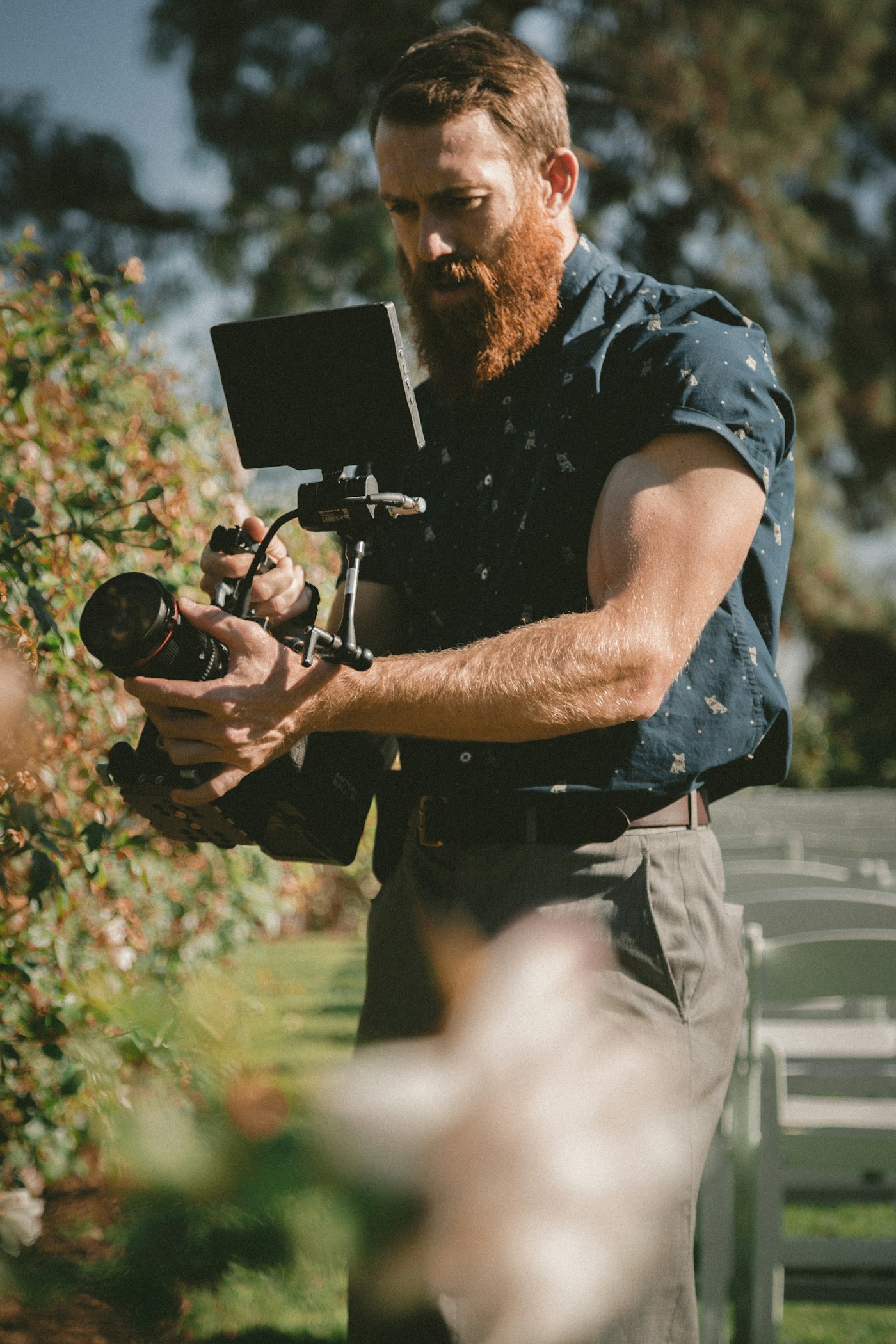 Photographer adjusting a camera rig outdoors during a wedding ceremony, surrounded by garden foliage