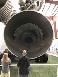 Two people stand in front of a massive rocket engine on display. The engine's large nozzle dominates the foreground, showcasing intricate details and mechanisms. The setting appears to be an indoor museum or exhibition space with lighting reflecting off the metal structure.