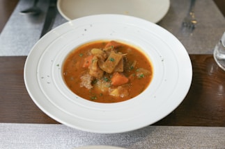 A white bowl contains a hearty stew with chunks of meat, carrots, and potatoes in a rich, red broth. The dish is garnished with finely chopped herbs. It is placed on a wooden table with a sleek, modern setting, including a fork and other tableware in the background.