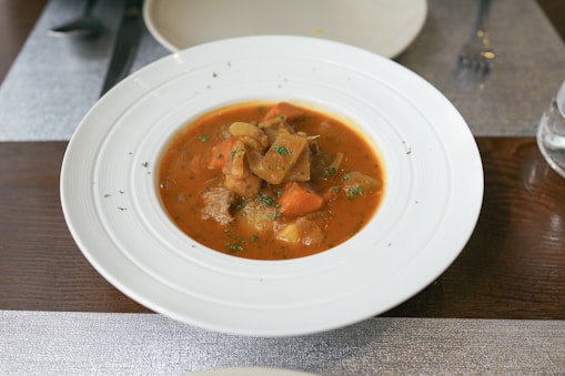 A white bowl contains a hearty stew with chunks of meat, carrots, and potatoes in a rich, red broth. The dish is garnished with finely chopped herbs. It is placed on a wooden table with a sleek, modern setting, including a fork and other tableware in the background.
