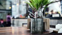 A rustic metal container holds a variety of silverware, including forks and spoons, alongside a small arrangement of green leaves. The container is placed on a wooden table in a cafe or restaurant setting. In the background, the interior is blurred, with hints of people, warm lighting, and an industrial decor.