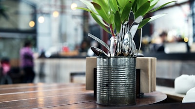 A rustic metal container holds a variety of silverware, including forks and spoons, alongside a small arrangement of green leaves. The container is placed on a wooden table in a cafe or restaurant setting. In the background, the interior is blurred, with hints of people, warm lighting, and an industrial decor.