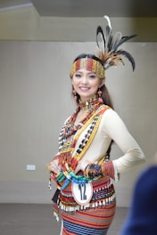 A person is wearing traditional attire adorned with colorful beads, feathers, and intricate patterns. The outfit includes a headdress with feathers, a necklace, and a woven belt. The expression is joyful and confident.