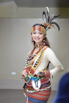 A person is wearing traditional attire adorned with colorful beads, feathers, and intricate patterns. The outfit includes a headdress with feathers, a necklace, and a woven belt. The expression is joyful and confident.