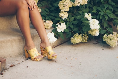 A pair of legs are seated on outdoor steps next to blooming white hydrangeas. The person is wearing yellow floral-printed platform sandals with a decorative anklet adorning the ankle. The setting has a relaxed and summery feel, with a concrete pavement and lush green leaves surrounding the flowers.