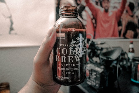 A hand holds a bottle of Starbucks Cold Brew Coffee, labeled with the mermaid logo and text indicating it is a premium coffee drink. The background is slightly blurred, showing a room with various objects including blurry images of people.