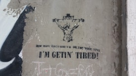 Graffiti on a wall depicting a figure on a cross, resembling a crucifixion scene, with the text 'How many times I have to die for your sins I'm gettin' tired!' The artwork is in black and white, with a worn and gritty texture surrounding it.