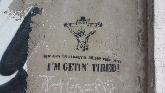 Graffiti on a wall depicting a figure on a cross, resembling a crucifixion scene, with the text 'How many times I have to die for your sins I'm gettin' tired!' The artwork is in black and white, with a worn and gritty texture surrounding it.