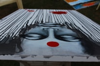 An upside-down painting depicts a serene face with closed eyes. The face is surrounded by cascading black lines that resemble rain or flowing hair, set against a pale background. A bold red dot is prominently featured on the forehead, adding a focal point of contrast.