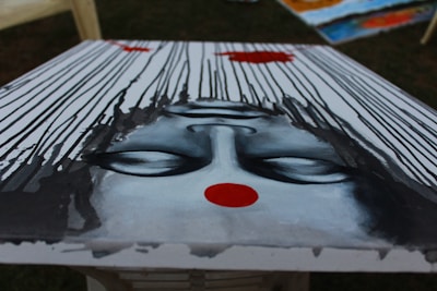 An upside-down painting depicts a serene face with closed eyes. The face is surrounded by cascading black lines that resemble rain or flowing hair, set against a pale background. A bold red dot is prominently featured on the forehead, adding a focal point of contrast.