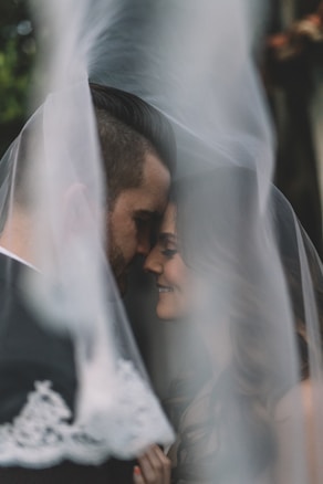 A couple is intimately close, their faces gently pressed together and partially obscured by a veil, creating a soft, romantic atmosphere.
