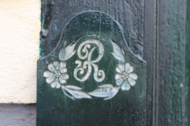 A decorative wooden panel featuring the letter 'R' surrounded by floral motifs. The wood appears weathered with a dark green paint, and the design is detailed with carved flowers and leaves in a light color.