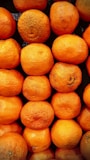 A collection of bright orange tangerines neatly organized in rows. The fruits have a textured surface and some natural blemishes, indicating freshness and ripeness. The background is a dark, contrasting color that highlights the vividness of the tangerines.