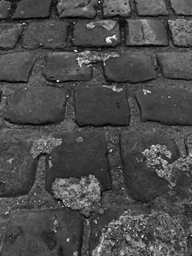 A close-up view of an old cobblestone street, featuring worn and weathered stones with patches of erosion and signs of wear. The stones are unevenly placed and have a rugged texture, indicating age and frequent use.