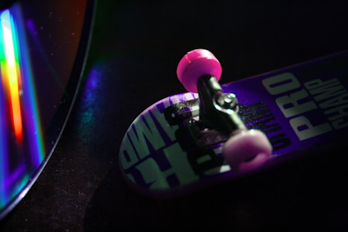 A close-up of a miniature skateboard with pink wheels and a deck featuring bold text, placed beside a CD that reflects a spectrum of vibrant colors. The scene is dimly lit, creating a moody atmosphere.
