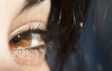 A close-up of a brown eye with long, dark eyelashes. The surrounding skin is visible, along with some strands of dark hair partially covering parts of the eye.