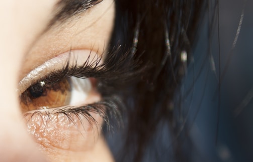 A close-up of a brown eye with long, dark eyelashes. The surrounding skin is visible, along with some strands of dark hair partially covering parts of the eye.