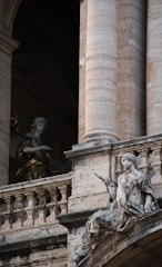 Two classical statues are positioned against a backdrop of grand architectural elements featuring stone columns and intricate stonework. The statue in the foreground is seated, holding what appears to be a scroll, while the other statue is partially in shadow, possibly depicting an angel. The intricate details of both the sculptures and the architecture suggest an atmosphere of historical and artistic significance.