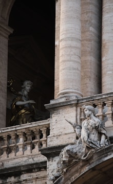 Two classical statues are positioned against a backdrop of grand architectural elements featuring stone columns and intricate stonework. The statue in the foreground is seated, holding what appears to be a scroll, while the other statue is partially in shadow, possibly depicting an angel. The intricate details of both the sculptures and the architecture suggest an atmosphere of historical and artistic significance.