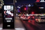 A well-lit urban street at night with a digital advertisement kiosk displaying a sneaker from the 'Foot Locker Approved. Black & Sand Collection.' Several cars are visible on the street, with illuminated store signs for BZ, SATURN, and KFC in the background.