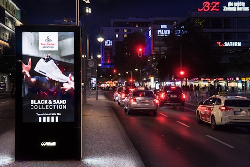 A well-lit urban street at night with a digital advertisement kiosk displaying a sneaker from the 'Foot Locker Approved. Black & Sand Collection.' Several cars are visible on the street, with illuminated store signs for BZ, SATURN, and KFC in the background.