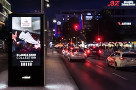 A well-lit urban street at night with a digital advertisement kiosk displaying a sneaker from the 'Foot Locker Approved. Black & Sand Collection.' Several cars are visible on the street, with illuminated store signs for BZ, SATURN, and KFC in the background.