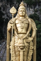 A large golden statue of a deity is prominently displayed in front of a rocky cliff with lush greenery. The statue holds a tall staff and is adorned with intricate jewelry and attire.
