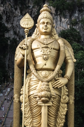 A large golden statue of a deity is prominently displayed in front of a rocky cliff with lush greenery. The statue holds a tall staff and is adorned with intricate jewelry and attire.
