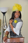 A woman is smiling and standing indoors, wearing a stylish black and white dress with flared sleeves. She accessorizes with a bright yellow hat adorned with feathers and a yellow floral brooch that match her bracelet and wristwatch. The background includes a lamp and a decorative mirror.