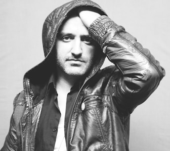 A black and white portrait of a person wearing a hooded leather jacket. The individual has one hand on their head and exhibits a thoughtful expression. The background is plain and light-colored, creating a strong contrast with the dark shades of the clothing.