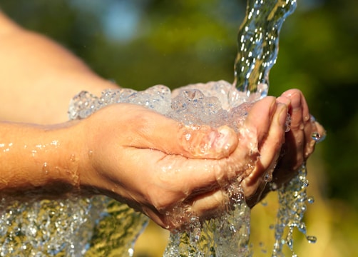 Hands cupped together catching fresh water as it flows over them against a soft-focus background of outdoor greenery. The sunlight glistens on the water droplets, highlighting the transparency and freshness.