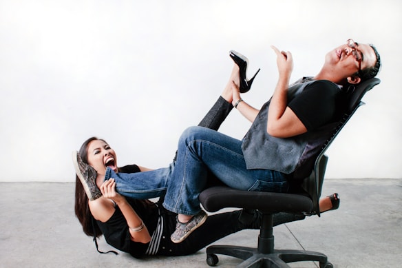 Two people are playfully interacting in an indoor setting, with one person sitting on a swiveling chair and the other on the floor. The person on the chair leans back, laughing, while the other holds onto the footrest, appearing to be in a humorous tug-of-war situation. Both individuals are casually dressed and seem to be enjoying the moment.