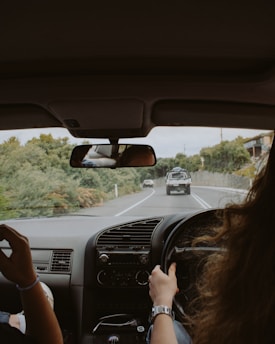 Inside a car, a person holds the steering wheel while driving on a road bordered by lush greenery. Through the windshield, another vehicle is visible ahead on the road. The car's interior reveals a dashboard with a ventilation panel and a rearview mirror reflecting part of the car's interior.