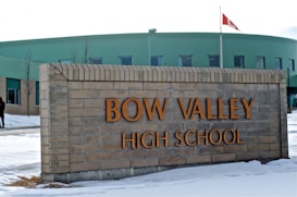 A large brick sign with the words 'BOW VALLEY HIGH SCHOOL' in prominent letters. Behind the sign, there is a modern building with a green exterior and several windows. A Canadian flag is visible on a pole to the right. The ground is covered with patches of snow, and a few bare trees can be seen. The overall setting suggests a winter day.