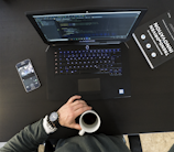 A workspace setting featuring an open laptop displaying code, a smartphone with a camera app open, a person's hand holding a cup of coffee, and a book titled 'Trend-Driven Innovation'. The person is wearing a watch and bracelets.