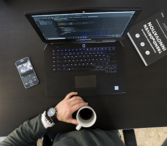 A workspace setting featuring an open laptop displaying code, a smartphone with a camera app open, a person's hand holding a cup of coffee, and a book titled 'Trend-Driven Innovation'. The person is wearing a watch and bracelets.