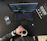 A workspace setting featuring an open laptop displaying code, a smartphone with a camera app open, a person's hand holding a cup of coffee, and a book titled 'Trend-Driven Innovation'. The person is wearing a watch and bracelets.