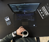 A workspace setting featuring an open laptop displaying code, a smartphone with a camera app open, a person's hand holding a cup of coffee, and a book titled 'Trend-Driven Innovation'. The person is wearing a watch and bracelets.