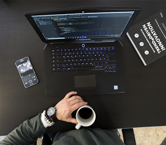 A workspace setting featuring an open laptop displaying code, a smartphone with a camera app open, a person's hand holding a cup of coffee, and a book titled 'Trend-Driven Innovation'. The person is wearing a watch and bracelets.